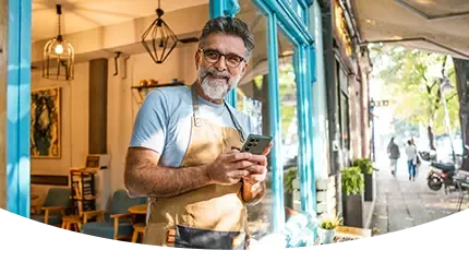 A smiling man in an apron holds a smartphone while standing at the entrance of a barber shop.